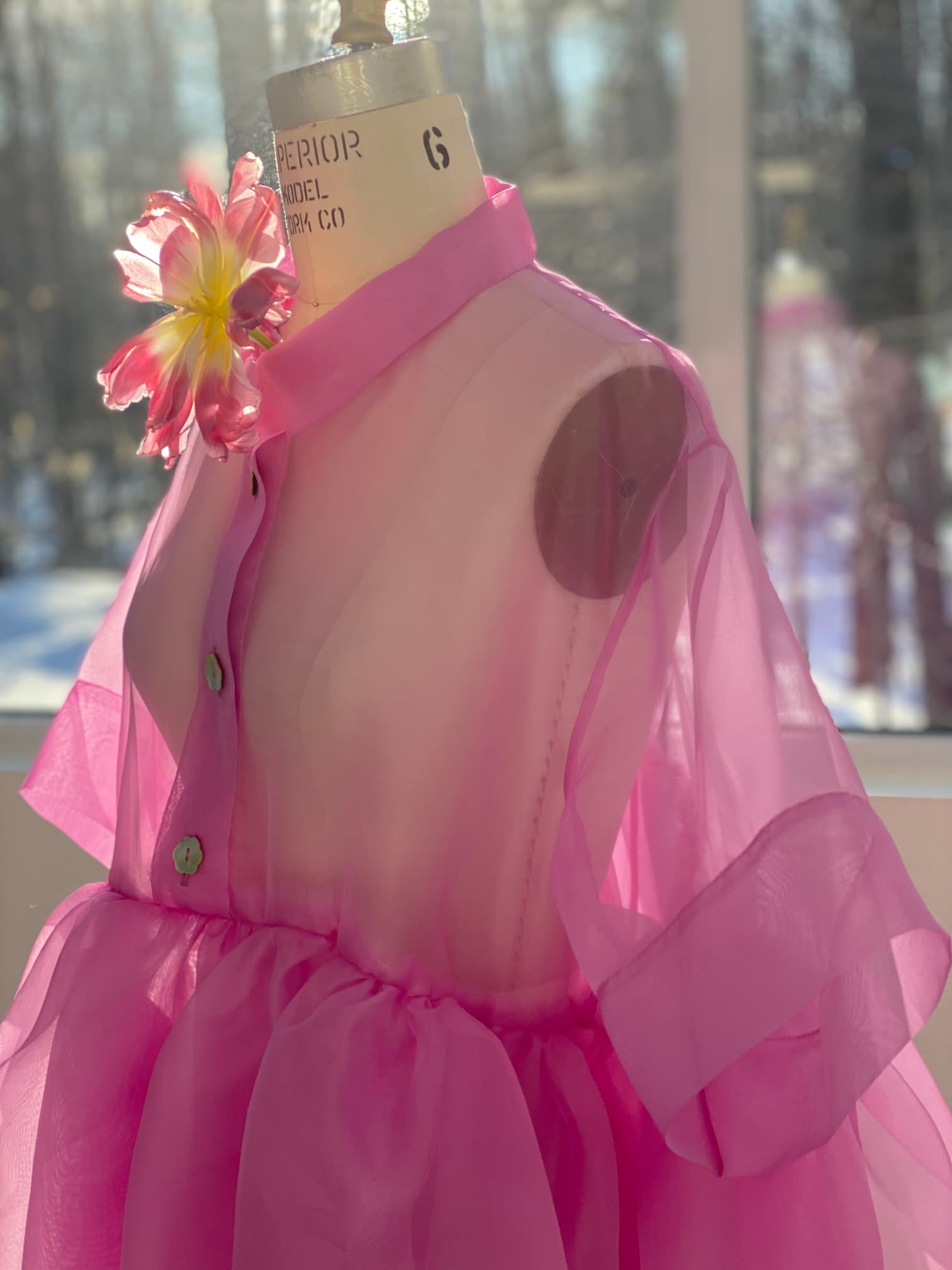 The Lou Shirt Dress by Noémiah, crafted from pink silk organza with a large flower embellishment on the shoulder, is displayed on a dress form. Sunlight streams through a window in the background, highlighting the fabric's translucent quality and relaxed fit.