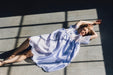 A woman in a flowing, relaxed-fit Lou Shirt Dress by Noémiah lies on a sunlit floor, framed by the shadows of window panes. She stretches her arms above her head, eyes closed, as light and shadow create geometric patterns around her.