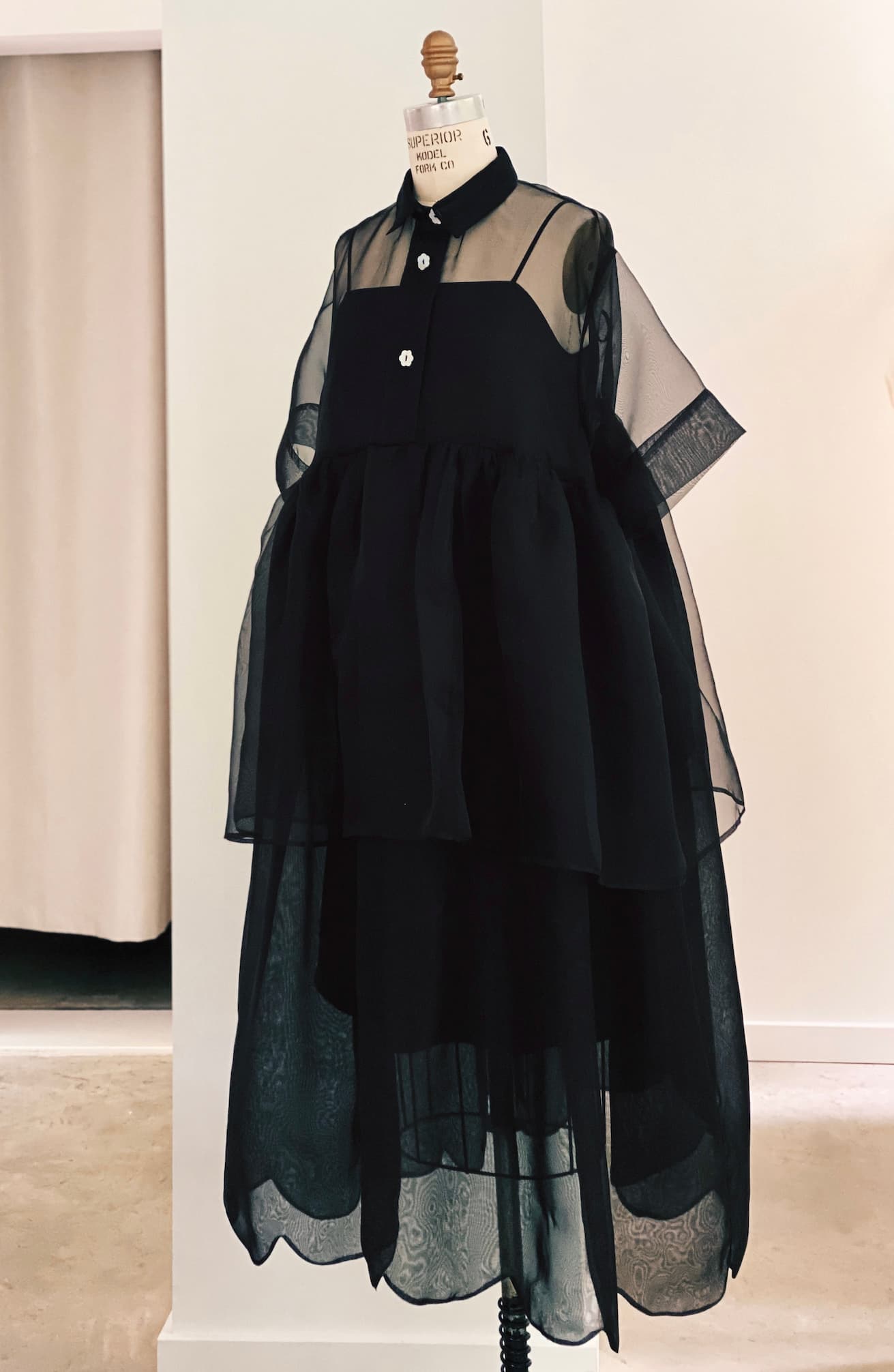 The Lou Shirt Dress by Noémiah is a black sheer oversized dress with voluminous layers and short sleeves, shown on a dress form in a minimalist room with light walls and curtains.