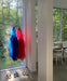 Two sheer Noémiah Lou Shirt Dresses—a blue silk organza and a red relaxed fit—hang by large windows facing a green forest. Below are two pairs of silver shoes, with a white chair and sewing machine in the background.
