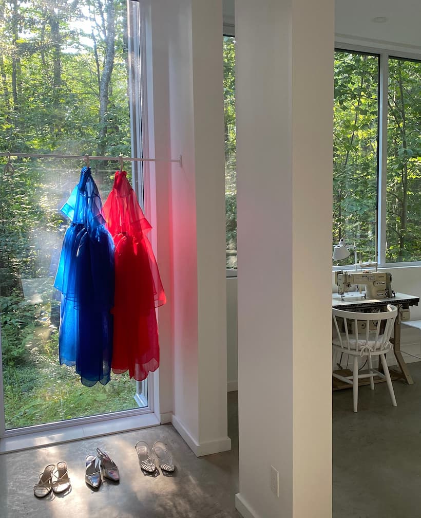 Two sheer Noémiah Lou Shirt Dresses—a blue silk organza and a red relaxed fit—hang by large windows facing a green forest. Below are two pairs of silver shoes, with a white chair and sewing machine in the background.