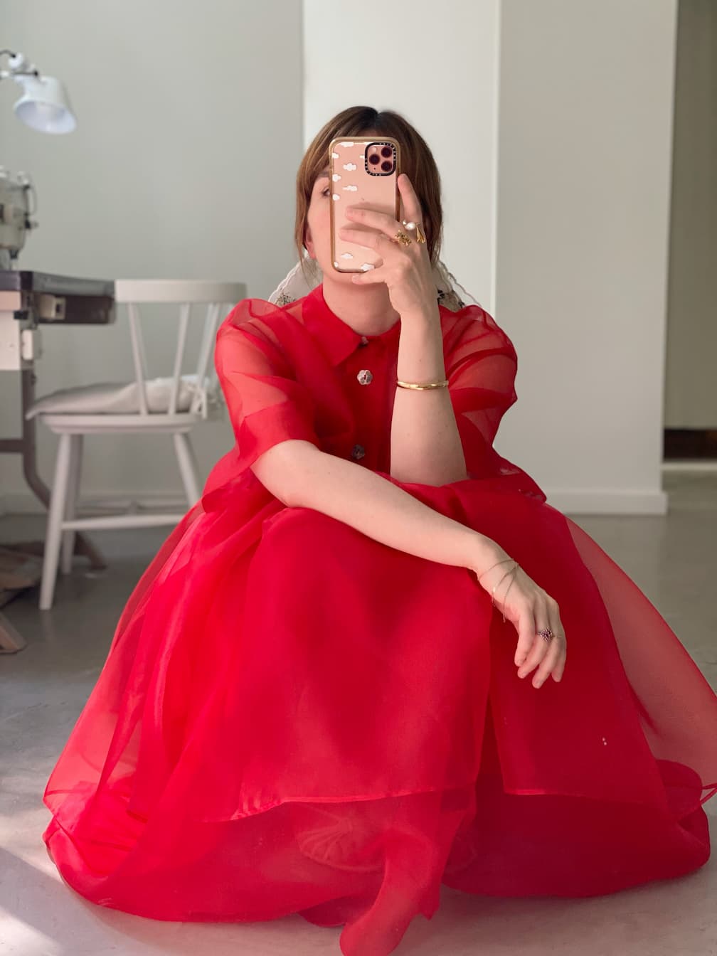A person wearing the Noémiah Lou Shirt Dress in bright red silk organza crouches on the floor, taking a mirror selfie with their phone. A white chair and a sewing machine appear in the background.
