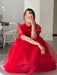 A person wearing the Noémiah Lou Shirt Dress in bright red silk organza crouches on the floor, taking a mirror selfie with their phone. A white chair and a sewing machine appear in the background.