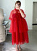 A person takes an indoor mirror selfie wearing the Lou Shirt Dress by Noémiah in a relaxed red silk organza style, paired with light sandals and gold rings. A white mannequin and chair can be seen in the background.