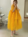 In a room filled with sewing equipment and neutral-toned floors and walls, a person takes a mirror selfie while wearing the Noémiah Lou Shirt Dress. The flowing, layered silk organza design in a vibrant yellow hue pairs perfectly with their slip-on shoes as they hold their phone.