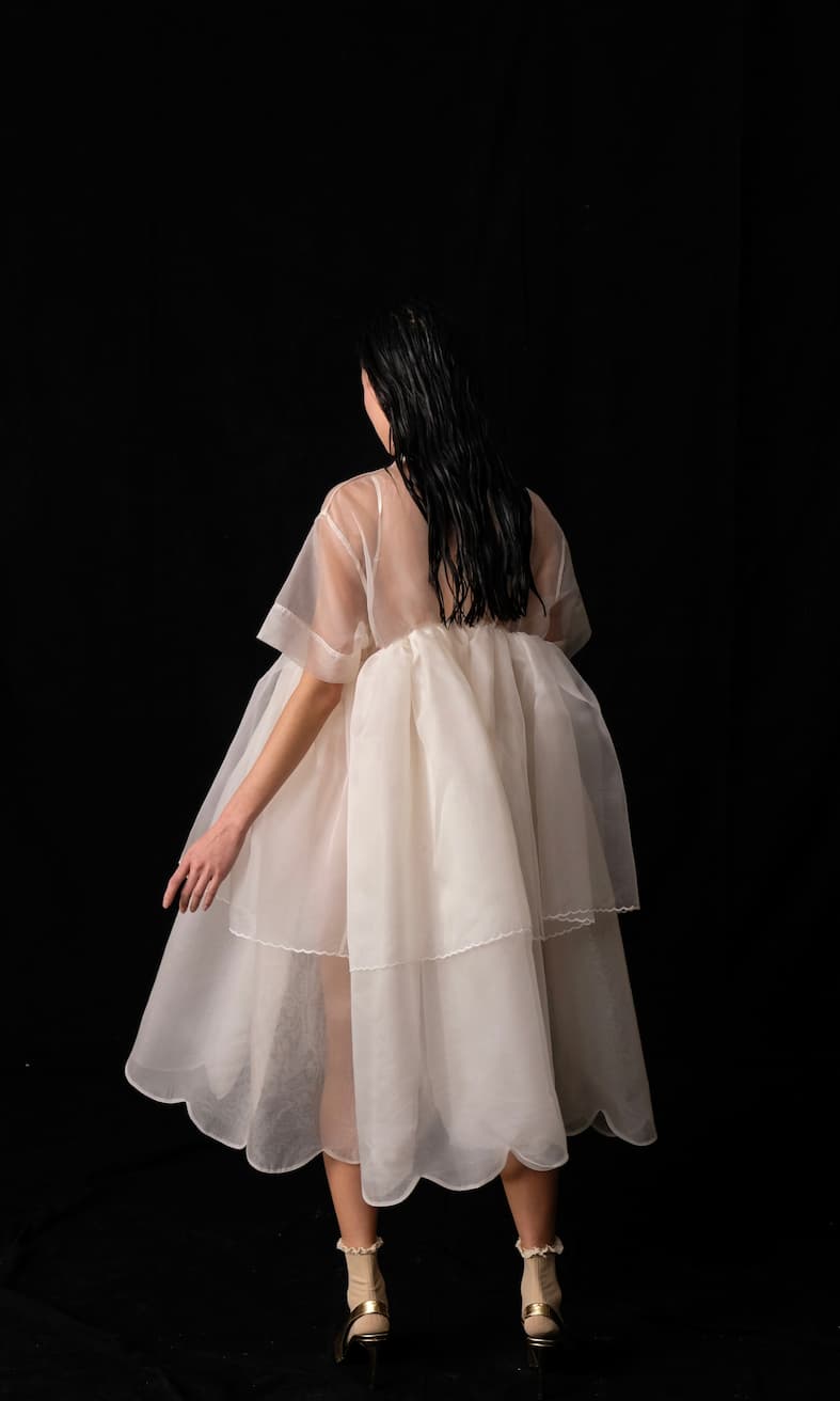 A person with long black hair stands against a black background, modeling the Noémiah Lou Shirt Dress — Embroidered. It's a layered, translucent white silk organza piece featuring scalloped edges and delicate porcelain buttons. The ensemble is completed with gold shoes, as they face away from the camera.