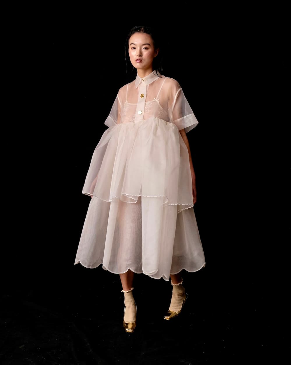 A woman stands against a black backdrop wearing the Lou Shirt Dress — Embroidered by Noémiah, an oversized piece crafted from sheer, layered silk organza. The outfit is accented with porcelain buttons and is elegantly paired with beige socks and gold shoes.