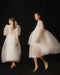 Two women stand against a dark background in flowing, translucent white Lou Shirt Dresses — Embroidered by Noémiah. The dresses are reminiscent of silk organza, and the soft focus of the image imparts a dreamy, ethereal quality. The woman on the left has red hair, while her counterpart on the right has dark hair.