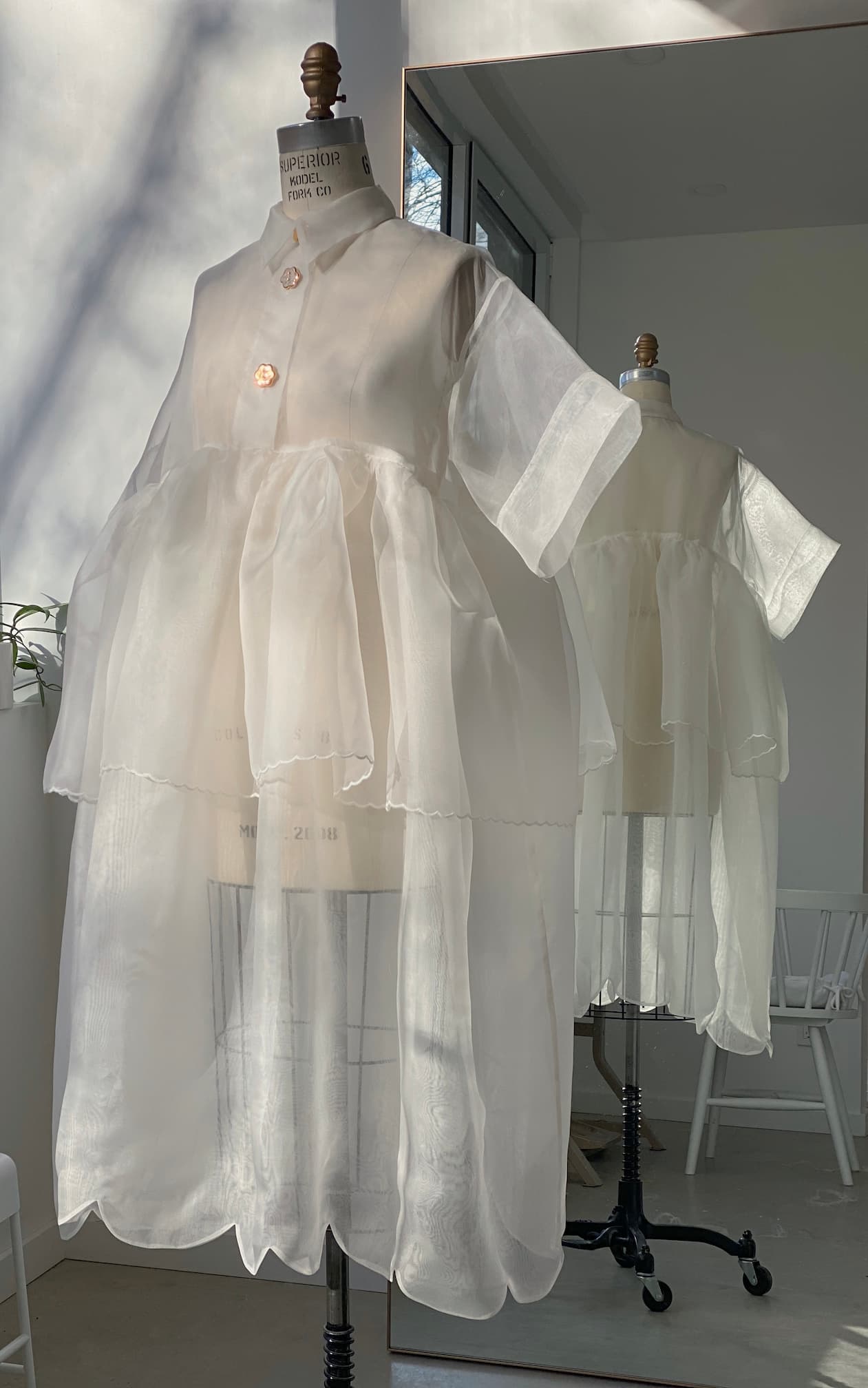 A translucent white Lou Shirt Dress — Embroidered by Noémiah is showcased on a mannequin, featuring short sleeves and ruffled layers with delicate porcelain buttons. The dress's reflection can be seen in a nearby mirror, while the room includes a white chair and a wall with a window streaming sunlight.