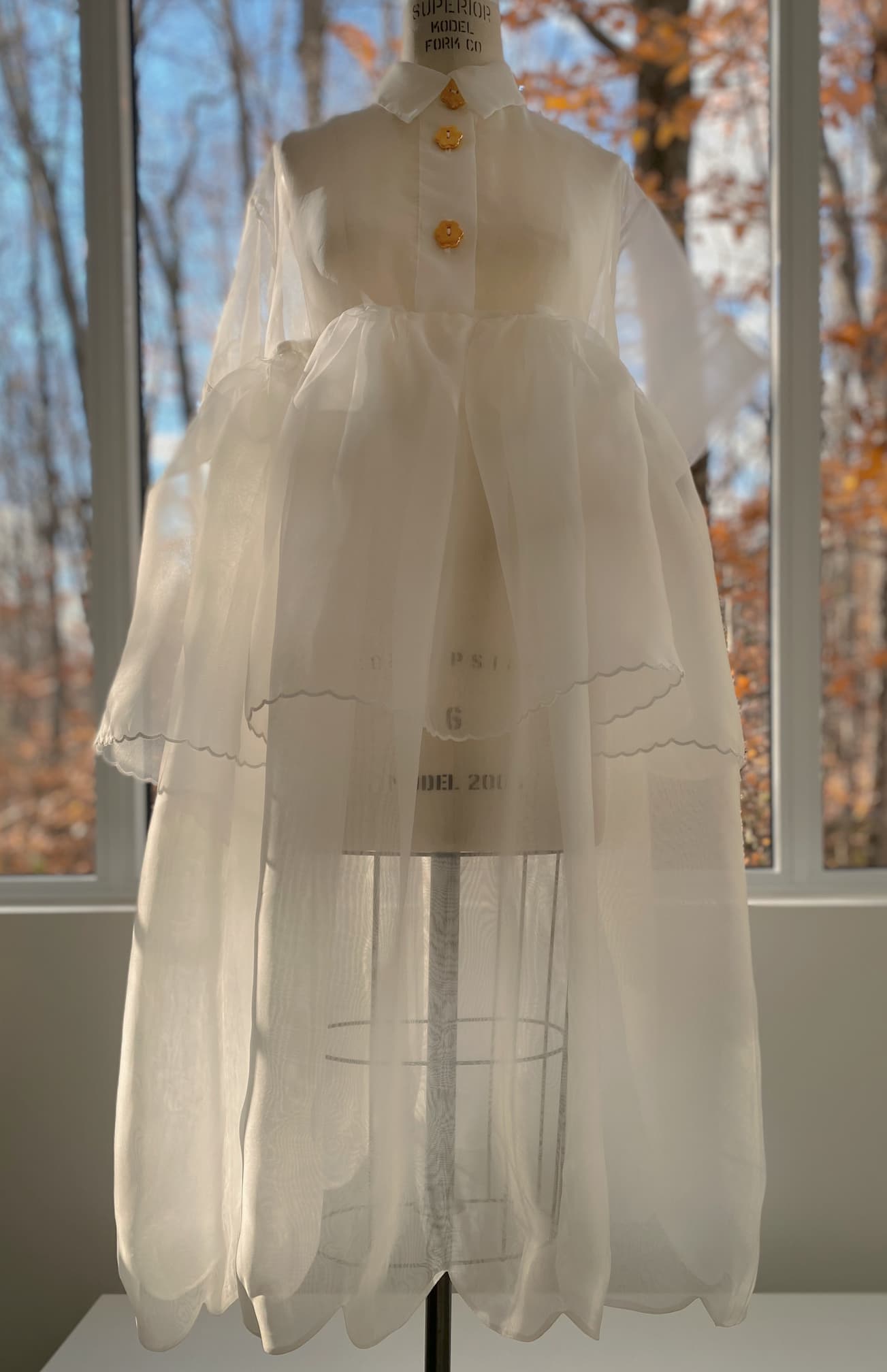 The mannequin showcases the Lou Shirt Dress — Embroidered by Noémiah, a white, sheer silk organza dress featuring a voluminous skirt and delicate porcelain buttons. Tall windows behind reveal autumn trees with sunlight streaming through.