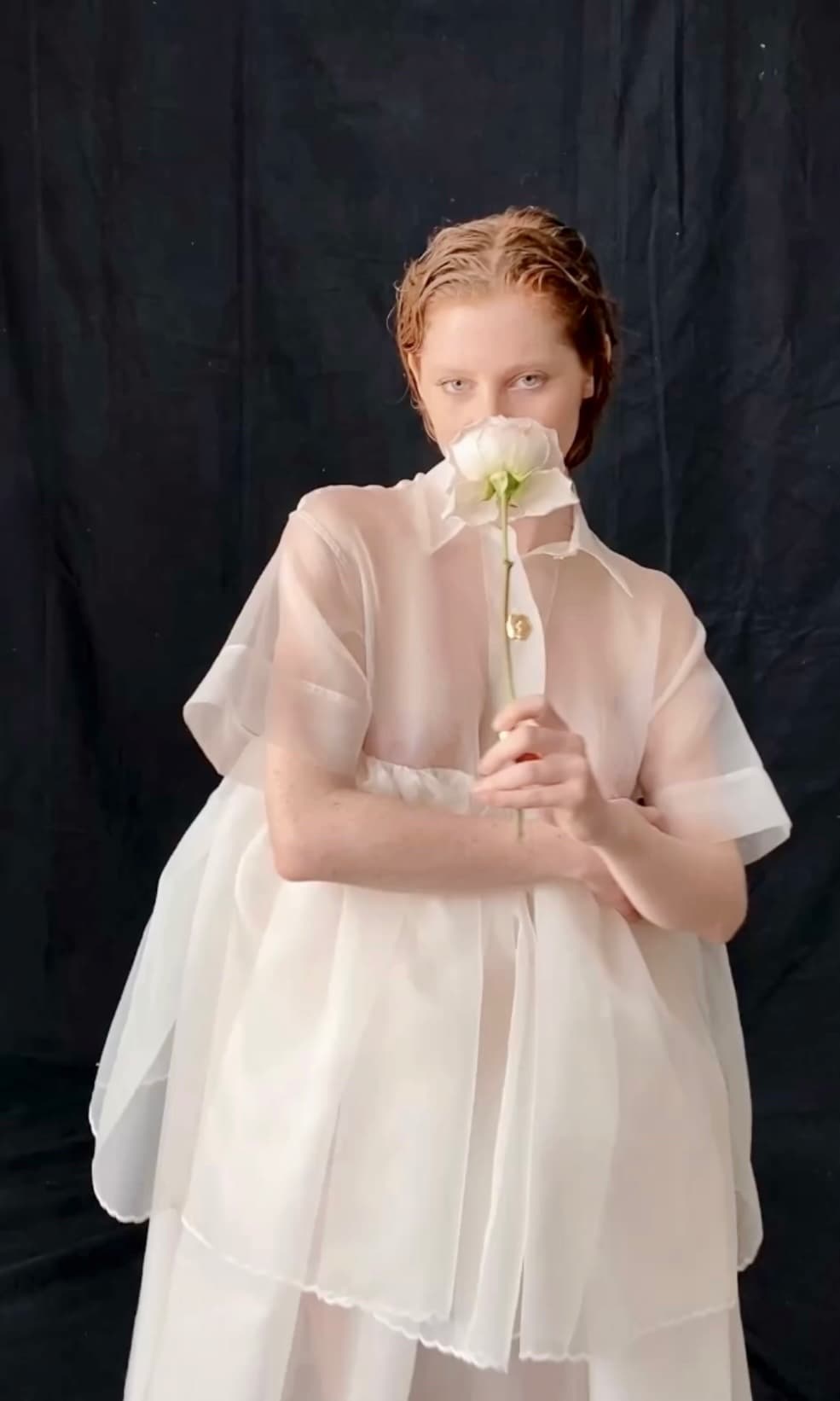 A person with short red hair stands against a dark background, draped in the Lou Shirt Dress — Embroidered by Noémiah. They hold a white flower close to their mouth, partially obscuring their face. The pose is elegant and contemplative.