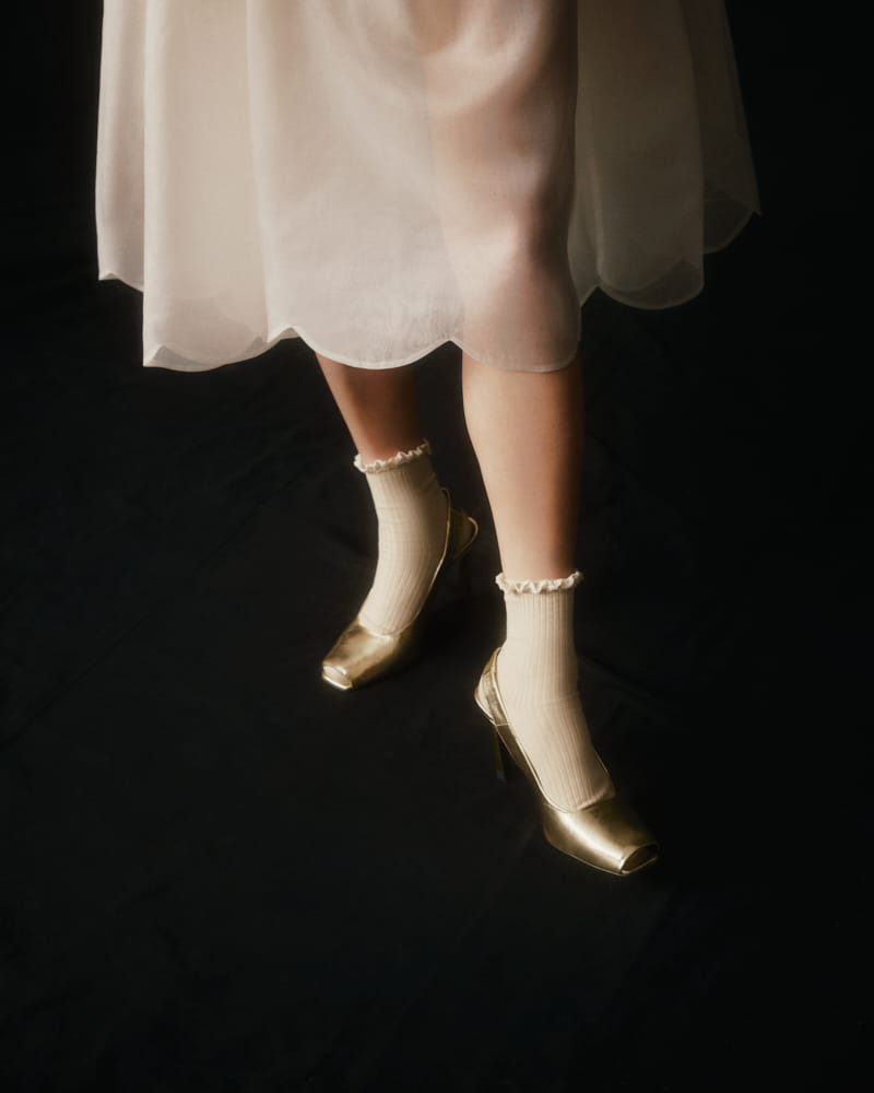 A person stands on a dark floor wearing metallic gold high-heeled shoes and cream-colored, ruffled ankle socks. They are dressed in the Lou Shirt Dress — Embroidered by Noémiah, a sheer, white oversized design crafted from silk organza with delicate porcelain buttons that add an extra touch of elegance.