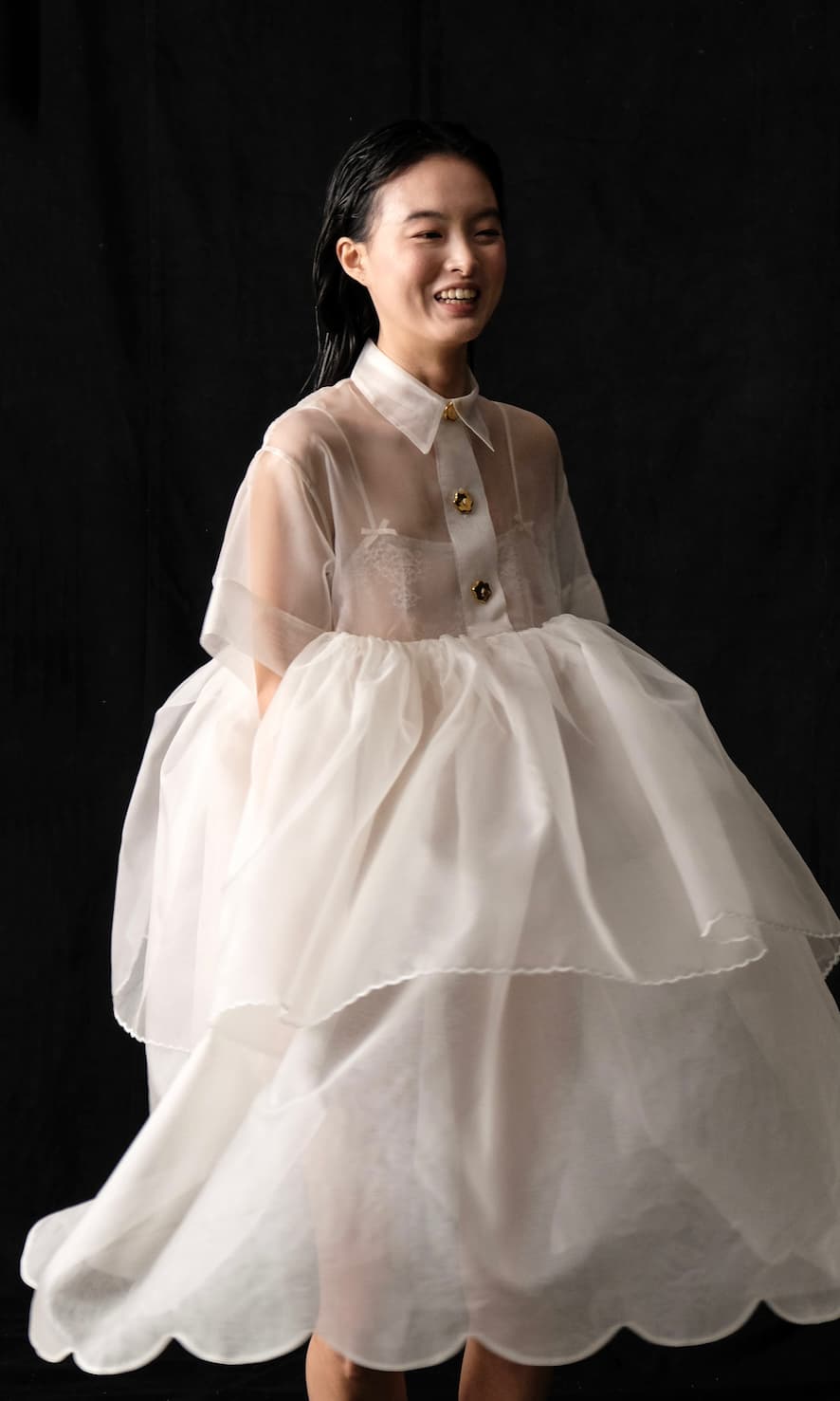 A person smiling while wearing the Lou Shirt Dress — Embroidered by Noémiah, a sheer and flowy white silk organza piece adorned with large layered ruffles and porcelain buttons. The dark background accentuates the dress's lightness.