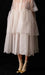 Against a dark background, a person showcases the Lou Shirt Dress — Embroidered by Noémiah. This oversized dress is made from sheer white silk organza and adorned with delicate porcelain buttons. It falls to knee-length and features scalloped edges, perfectly paired with cream socks and shimmering gold shoes.