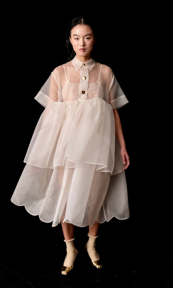 Against a black background, a person stands wearing the Lou Shirt Dress — Embroidered by Noémiah, featuring sheer silk organza with a collared neckline and porcelain buttons, resembling an oversized shirt dress, paired with gold shoes and beige socks.