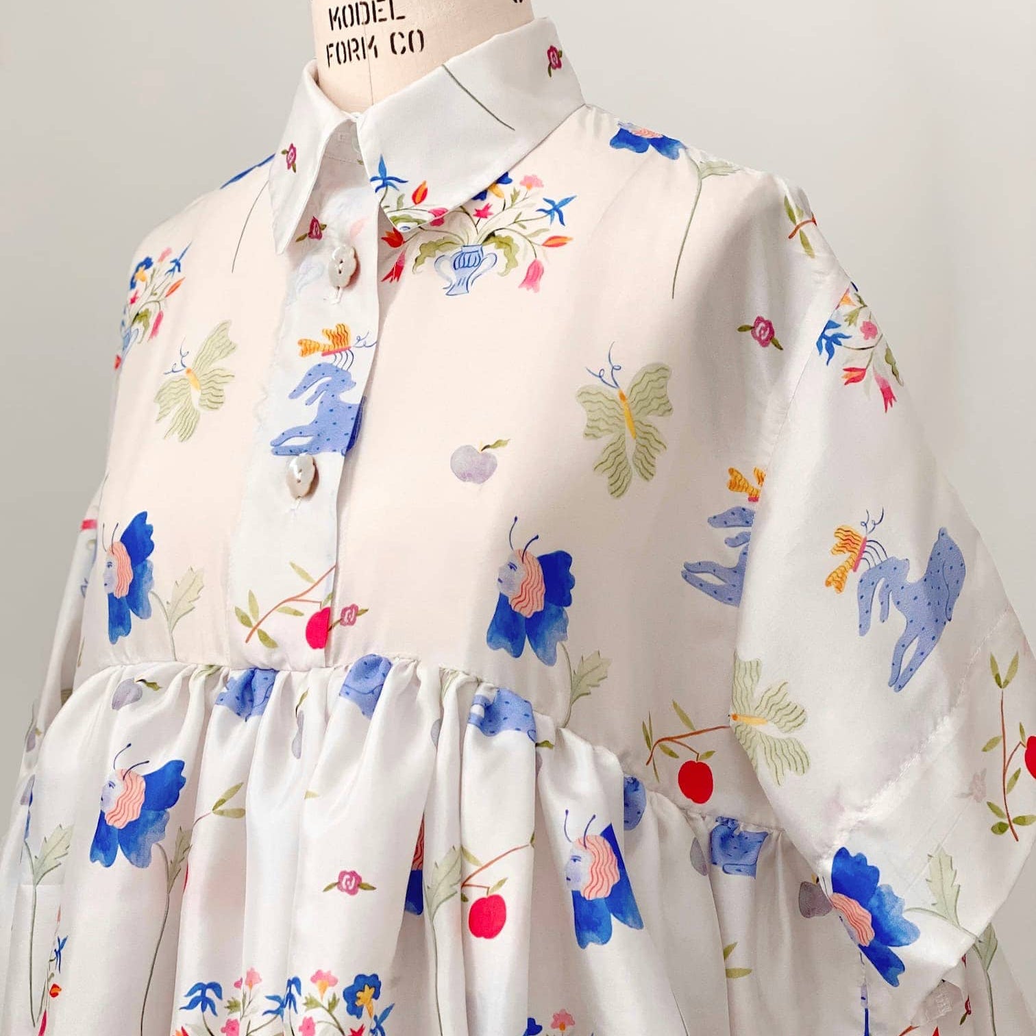 The Lou Shirt Dress — Printed by Noémiah is displayed on a mannequin, highlighting its vibrant patterned silk featuring flowers, butterflies, and abstract shapes. This collaboration with artist Estée Preda offers a button-up front and a loose, flowing silhouette for a whimsical and artistic look.