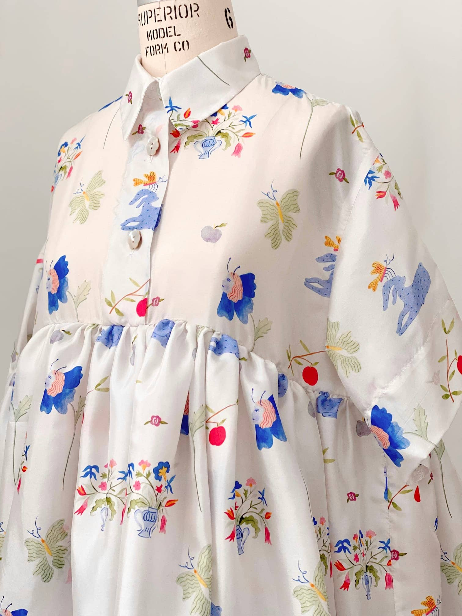 The Lou Shirt Dress — Printed by Noémiah is displayed on a mannequin, highlighting its vibrant patterned silk featuring flowers, butterflies, and abstract shapes. This collaboration with artist Estée Preda offers a button-up front and a loose, flowing silhouette for a whimsical and artistic look.