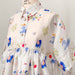 The Lou Shirt Dress — Printed by Noémiah is displayed on a mannequin, highlighting its vibrant patterned silk featuring flowers, butterflies, and abstract shapes. This collaboration with artist Estée Preda offers a button-up front and a loose, flowing silhouette for a whimsical and artistic look.