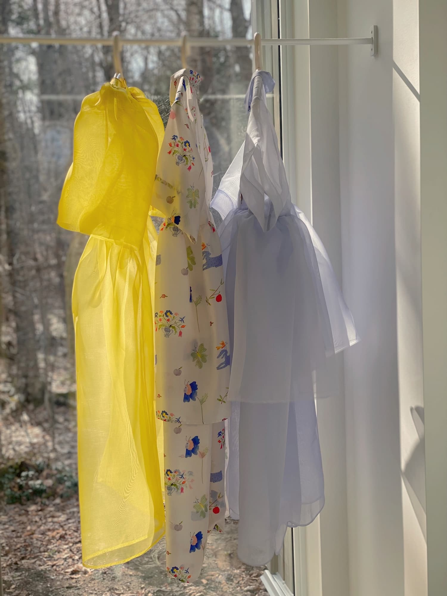 Three delicate dresses, featuring the Lou Shirt Dress — Printed from Noémiah's collaboration with artist Estée Preda, hang in front of a sunlit window. The dresses are in bright yellow, floral pattern, and light blue. Sunlight casts soft shadows through the sheer fabrics, and trees are visible outside.