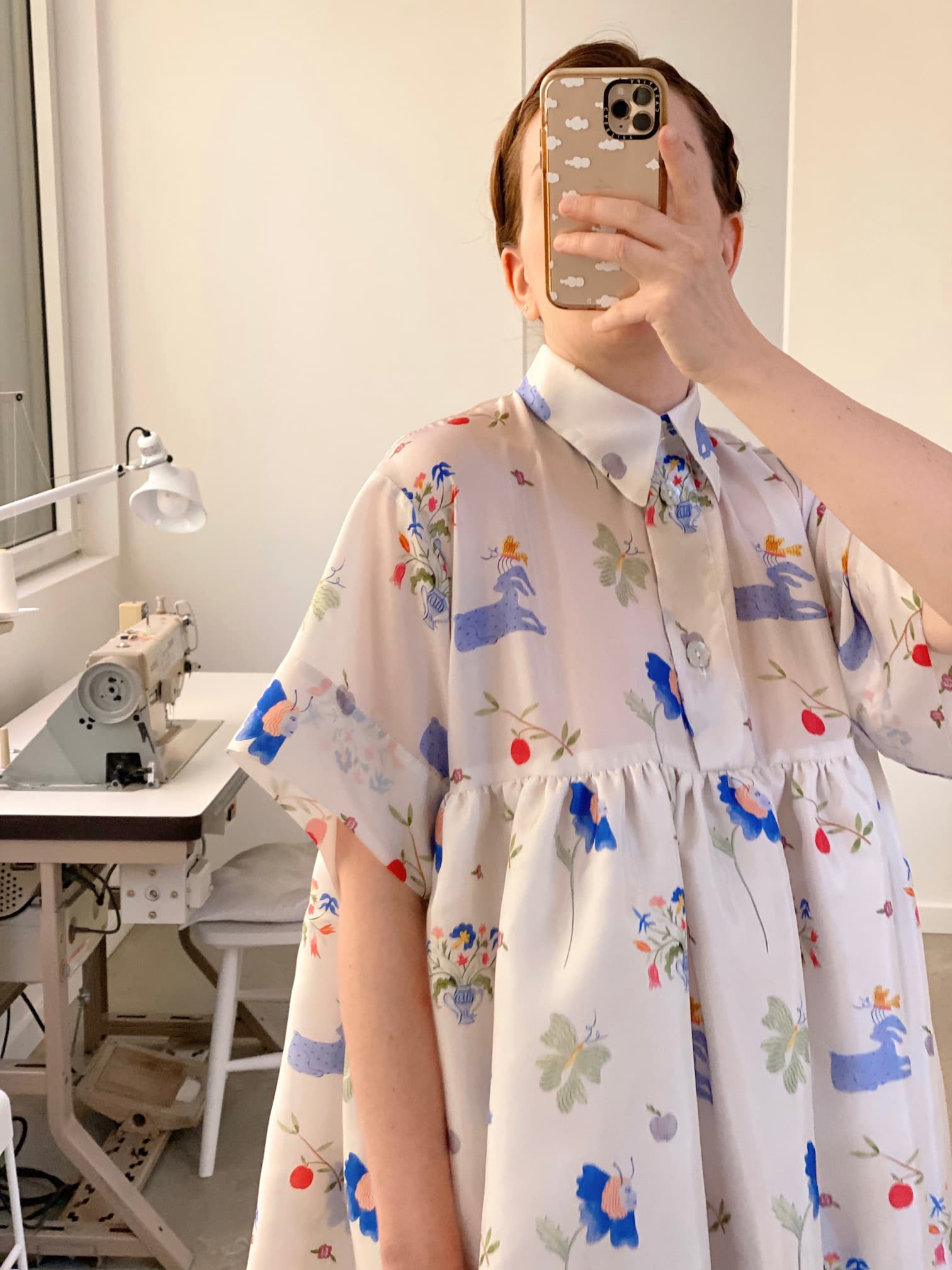 In a mirror selfie, someone wears the Lou Shirt Dress — Printed from Noémiah, showcasing its vibrant floral and animal patterns that suggest a collaboration with artist Estée Preda. A sewing machine and lamp can be seen in the background, indicating a crafting space.