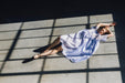 A woman in a flowing, relaxed-fit Lou Shirt Dress by Noémiah lies on a sunlit floor, framed by the shadows of window panes. She stretches her arms above her head, eyes closed, as light and shadow create geometric patterns around her.