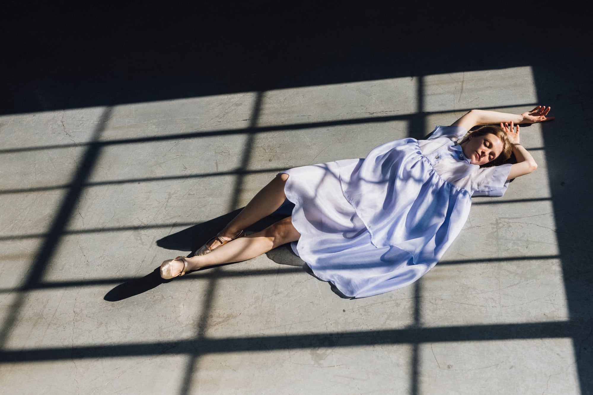 A woman in a flowing, relaxed-fit Lou Shirt Dress by Noémiah lies on a sunlit floor, framed by the shadows of window panes. She stretches her arms above her head, eyes closed, as light and shadow create geometric patterns around her.