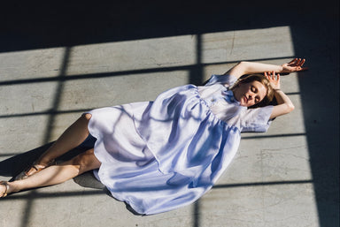 A woman in a flowing, relaxed-fit Lou Shirt Dress by Noémiah lies on a sunlit floor, framed by the shadows of window panes. She stretches her arms above her head, eyes closed, as light and shadow create geometric patterns around her.