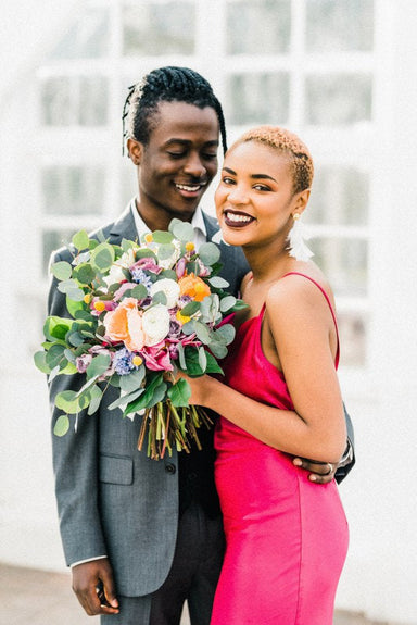 A smiling couple embraces, the woman holding a vibrant bouquet of flowers. She wears a bright pink dress complemented by Louise Earrings from Noémiah, while he is dressed in a gray suit. They are standing in front of a bright, windowed background.