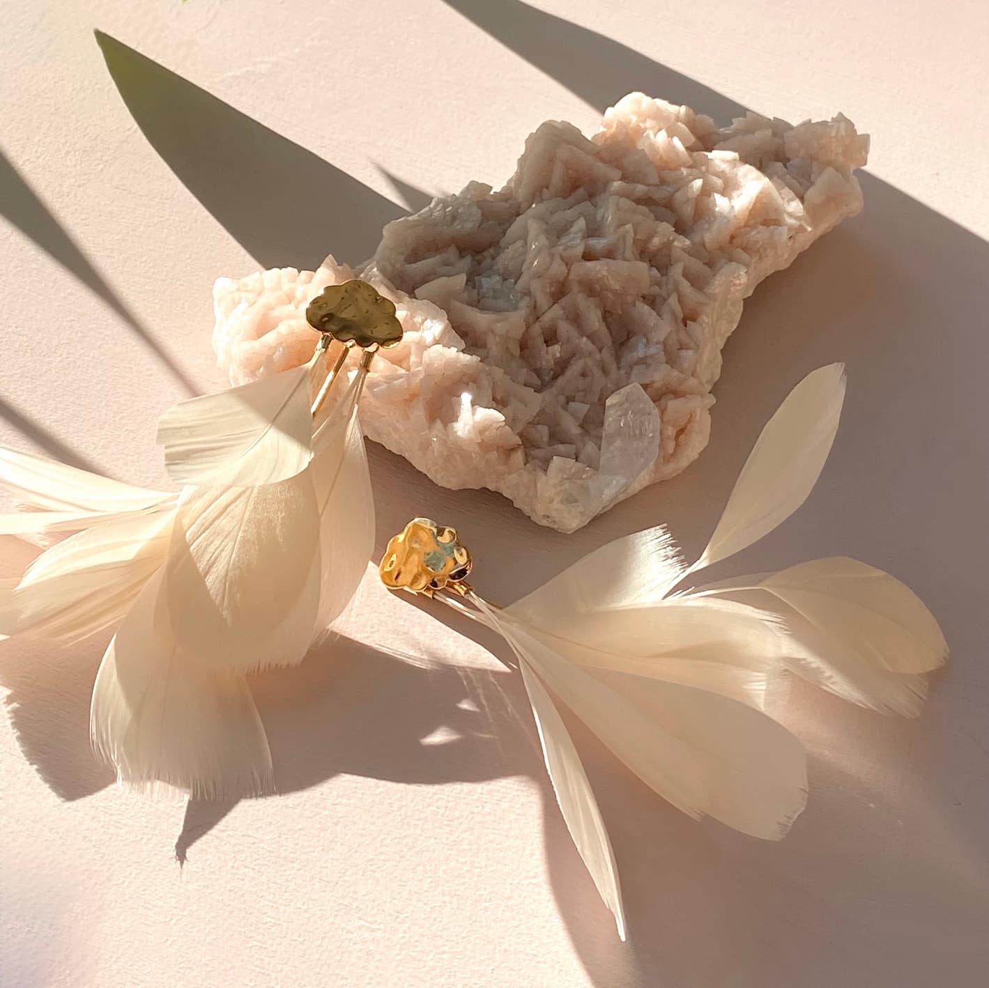 Two Louise Earrings from Noémiah, featuring natural feathers and gold accents in a cream hue, rest on a light surface beside a textured cream mineral rock. The scene is bathed in warm sunlight, casting soft shadows.