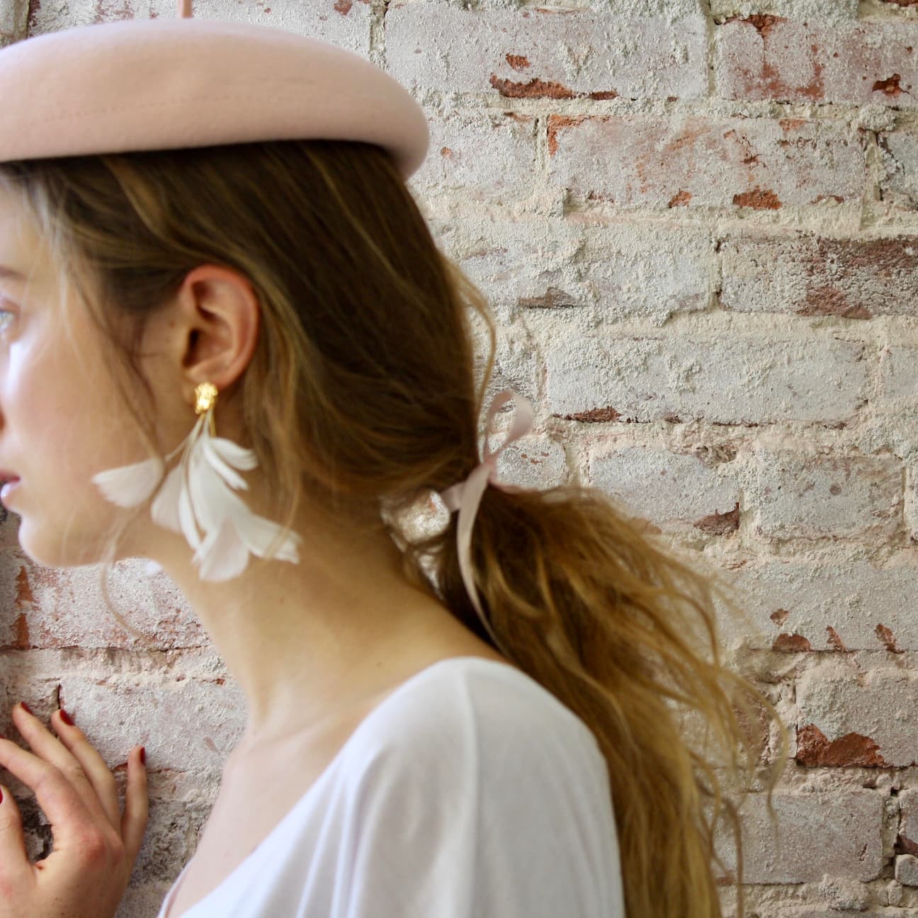 A woman with light brown hair is adorned in a pink beret and Louise Earrings by Noémiah, facing a textured brick wall. She has a ribbon in her hair and wears a white top. Her hand, with painted nails, gently touches the wall, while feather-like adornments complement her look.