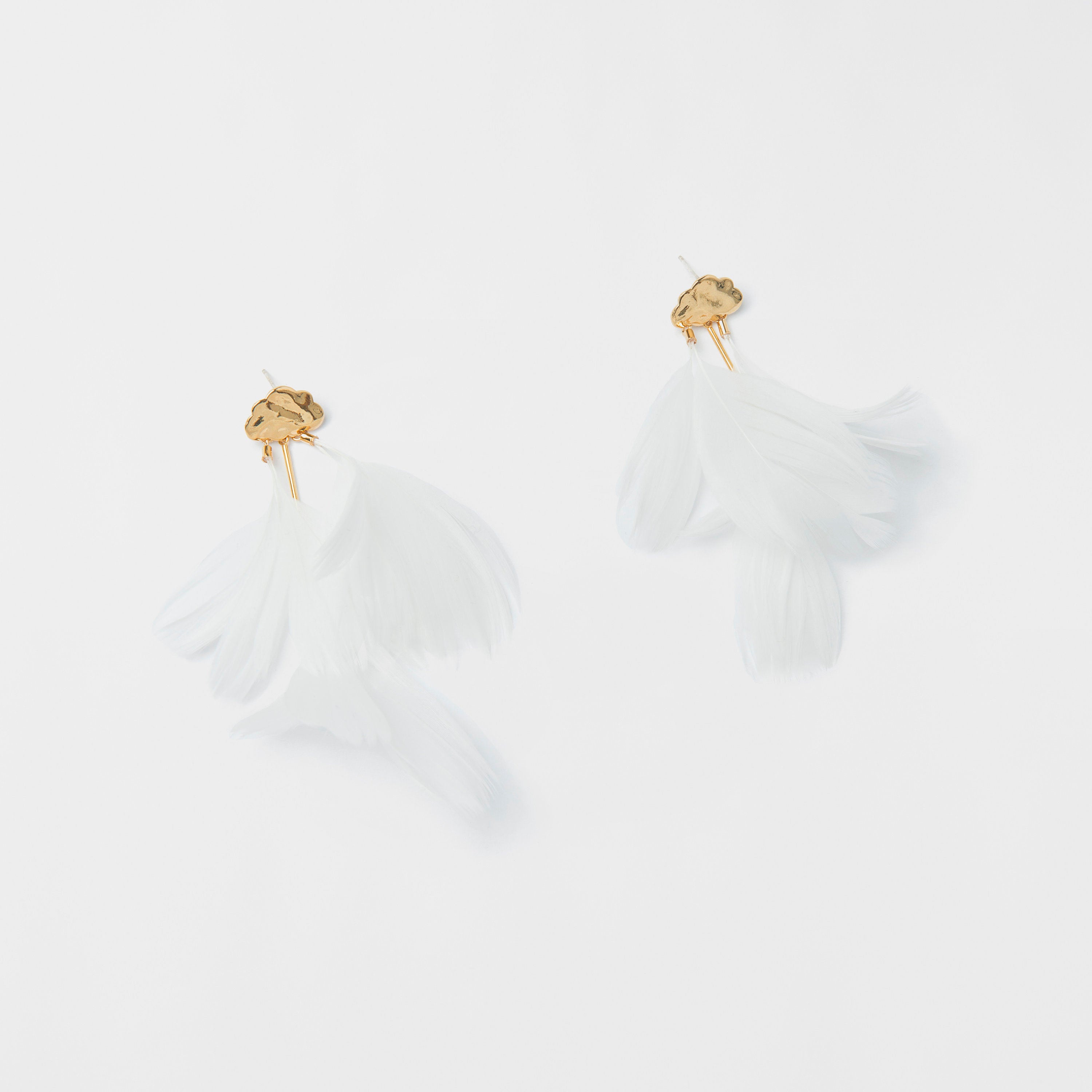 The Louise Earrings by Noémiah are elegantly presented against a white background, showcasing natural feathers with gold-tone floral accents. These cloud-like earrings boast a delicate, airy aesthetic as the feathers fan out gracefully, all crafted with durable chirurgical steel for enduring beauty.