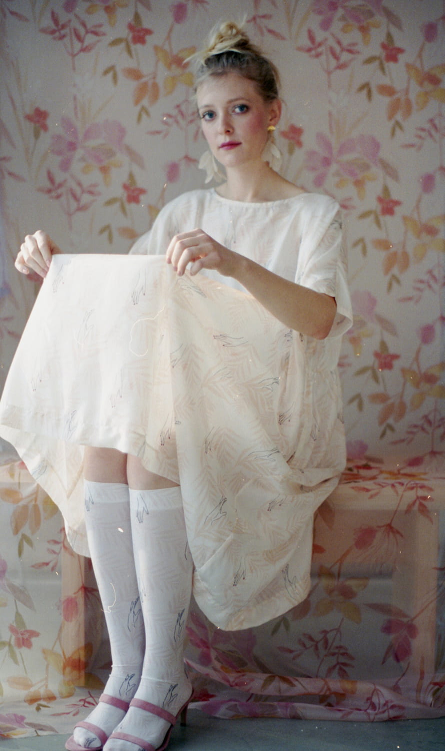 A woman with blonde hair styled in a bun sits on a floral-patterned background, wearing the delicate Louise Earrings by Noémiah. She dons a soft, flowing white dress and knee-high patterned socks, looking thoughtfully into the distance.