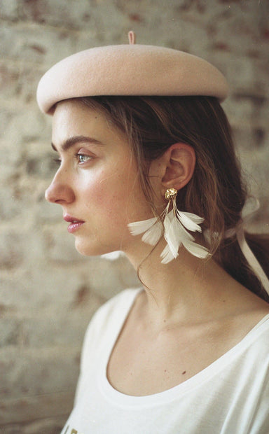 A woman in profile wears a light pink beret and Louise Earrings by Noémiah, featuring natural feather accents. Her hair is tied back with a ribbon that complements her white top, while the softly blurred brick wall in the background enhances her chic look with subtle charm.