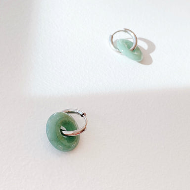 The Noémiah Lucie Earrings, featuring two small silver hoops each adorned with a pendant resembling green jade and boasting tarnish-resistant plating, are set against a textured white background. These elegant earrings create gentle shadows, enhanced by the lighting to evoke a serene and minimalist ambiance.