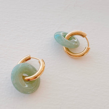 Introducing the Lucie Earrings by Noémiah, a stunning pair featuring gold hoops interwoven with jade-inspired light green circular elements. Their tarnish-resistant plating ensures they retain their radiant charm when placed on any light-colored surface.