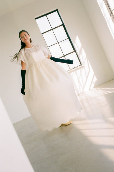 A woman wearing the Lula Dress by Noémiah, made of silk satin organza with a voluminous gathered skirt, and paired with black gloves, stands in a sunlit room with large windows. She appears to be in motion, exuding elegance and grace within the bright, minimalist space.