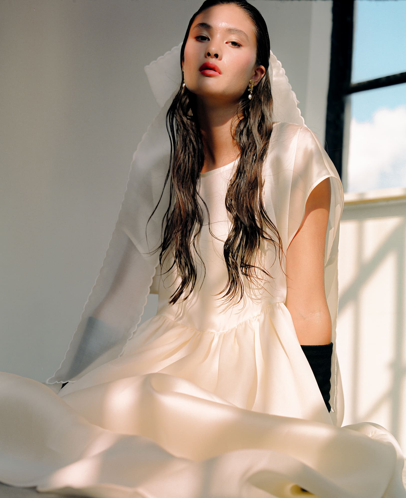 A woman in the elegant Lula Dress by Noémiah sits gracefully by a window, soft light illuminating her. Her long, wet-look hair and sheer veil complement the dress's relaxed fit. Her expression is serene and composed.