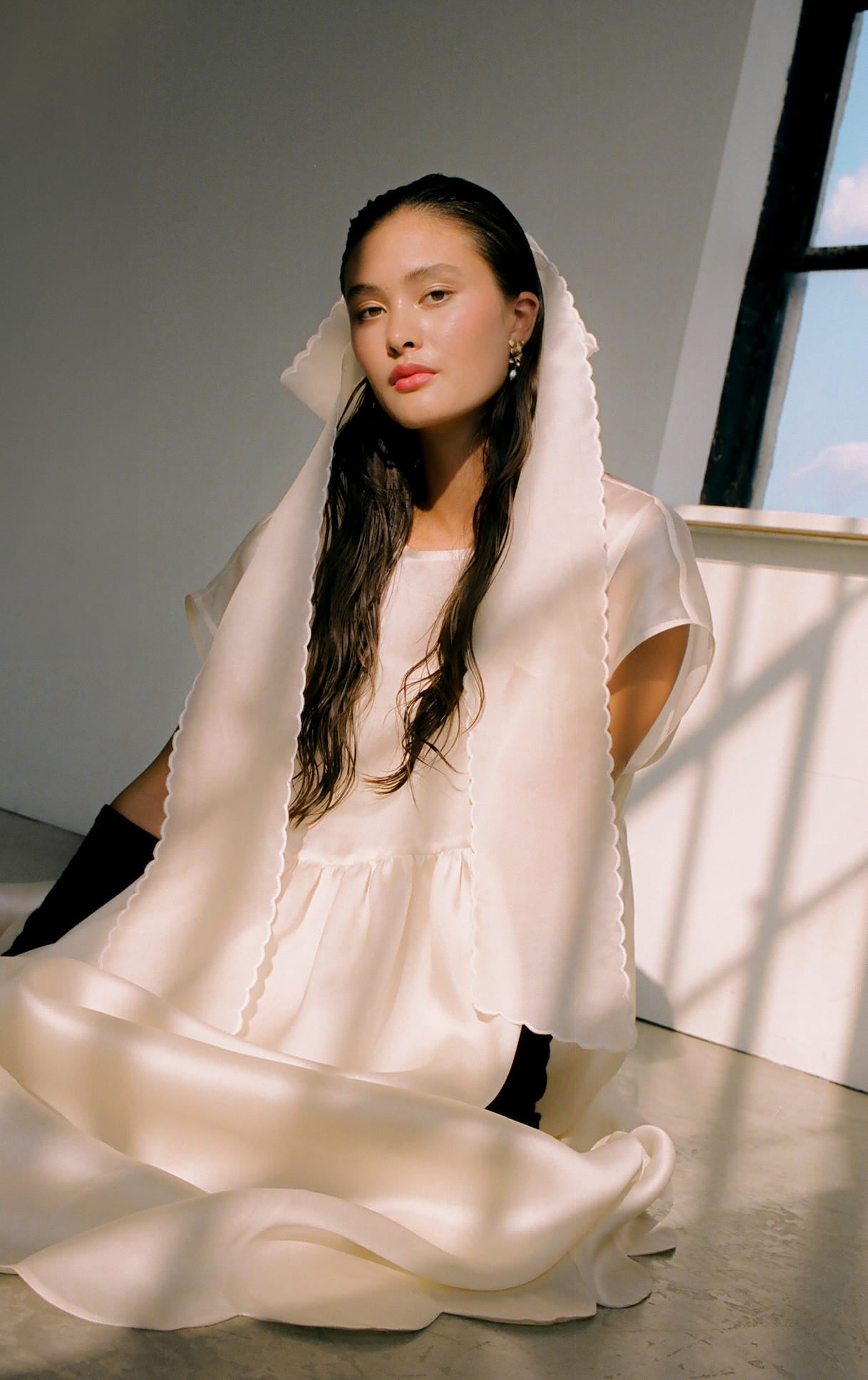 A woman wearing the Lula Dress by Noémiah sits gracefully on a sunlit floor with soft shadows. The dress features a flowing white design with a voluminous gathered skirt. She wears a silk satin organza veil over her dark, wavy hair. A large window in the background reveals a blue sky with clouds, adding to the serene and contemplative atmosphere.