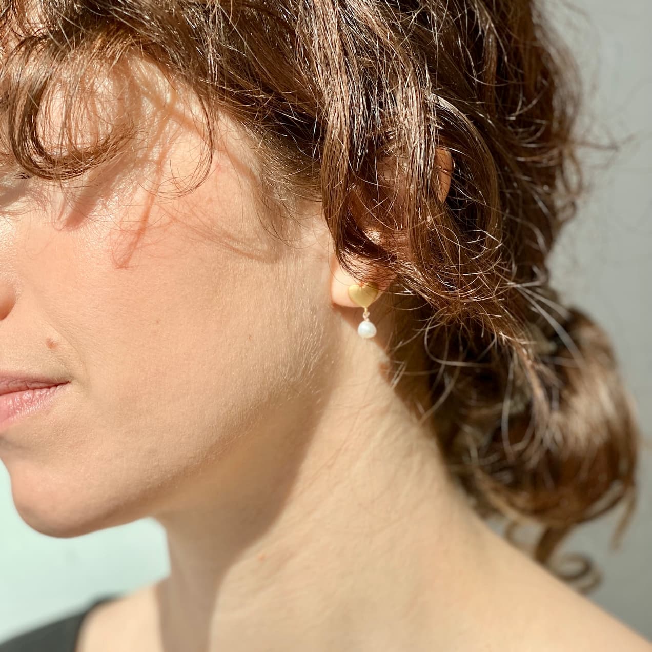 A close-up captures a person's brown hair neatly tied back, highlighting the Madeleine Earrings by Noémiah. These earrings feature freshwater pearls and are set in matte gold plating that adds an elegant touch. The individual faces left, bathed in sunlight that casts a soft glow on their skin, with the background gently out of focus.