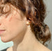 A close-up captures a person's brown hair neatly tied back, highlighting the Madeleine Earrings by Noémiah. These earrings feature freshwater pearls and are set in matte gold plating that adds an elegant touch. The individual faces left, bathed in sunlight that casts a soft glow on their skin, with the background gently out of focus.