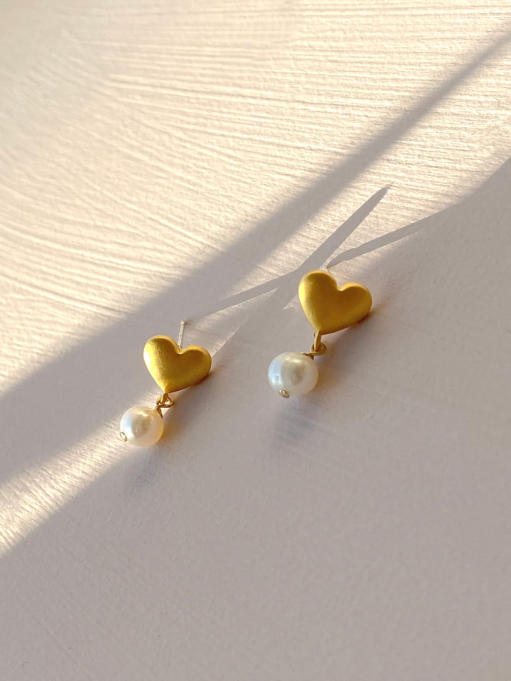 The Madeleine Earrings by Noémiah showcase heart-shaped studs in matte gold plating with elegant dangling freshwater pearls, beautifully displayed on a light surface with soft shadows.