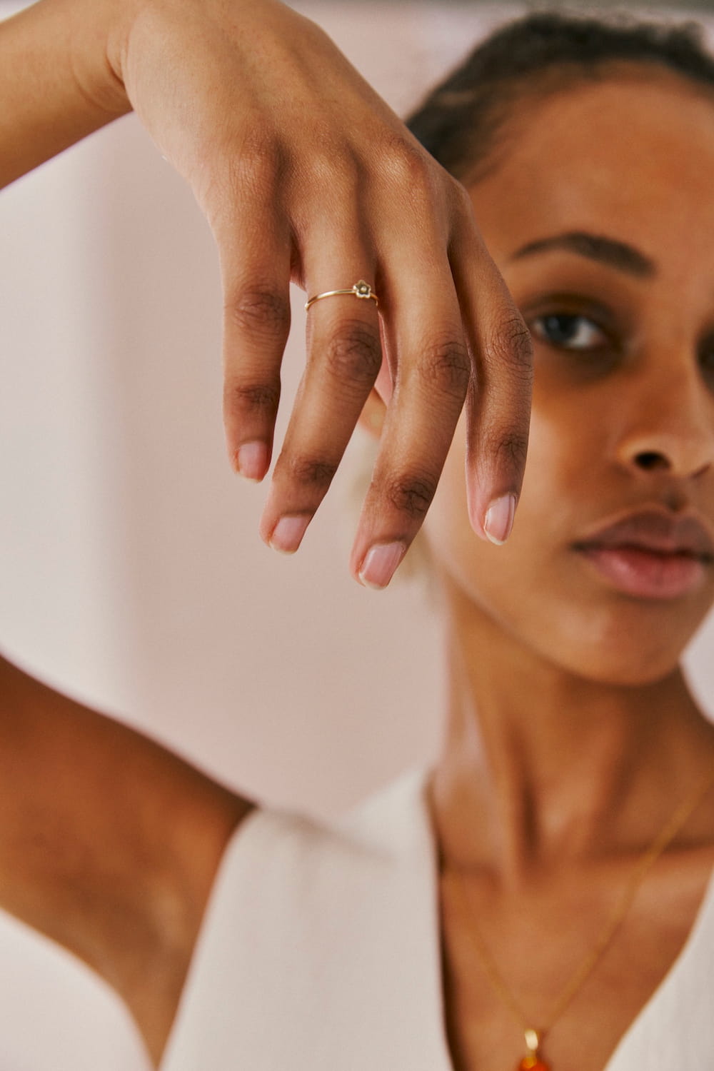 A person with a calm expression poses, showcasing the delicate handmade Marguerite Ring by Noémiah on their hand. They wear a simple solid gold necklace and a white top, set against a neutral, blurred background.