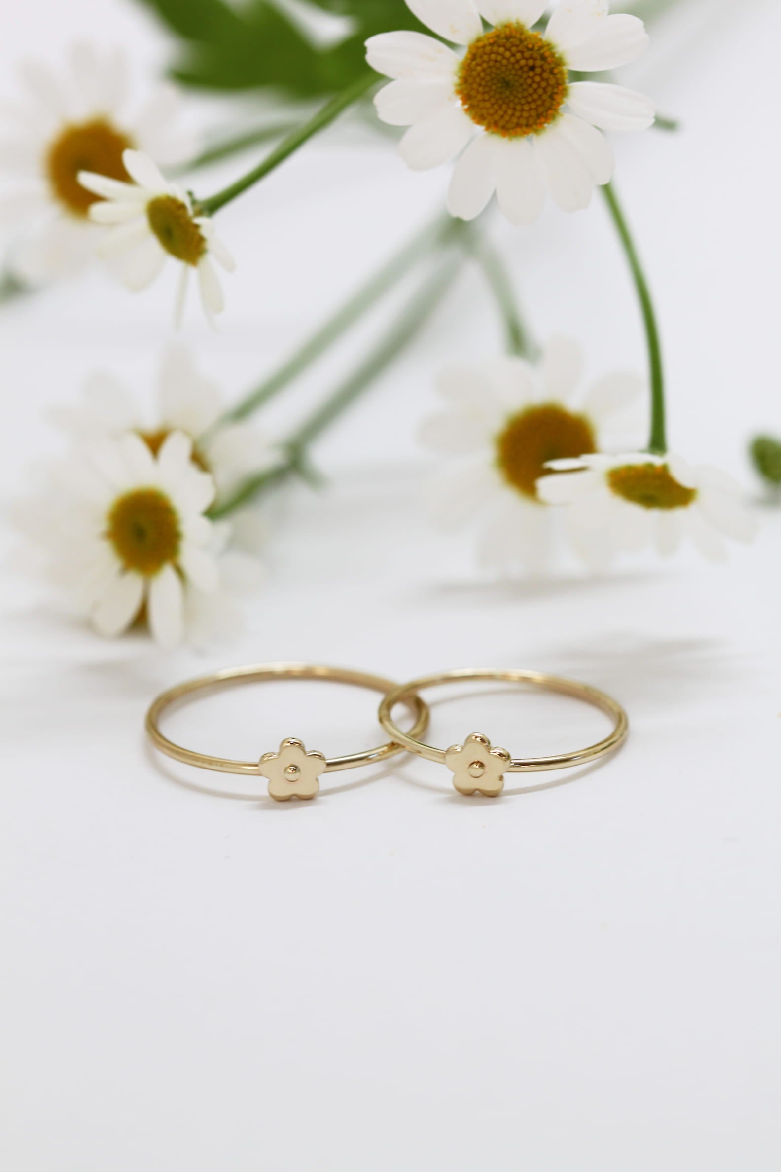 A pair of delicate Marguerite Rings from Noémiah, crafted in solid gold with intricate small flower designs, rest gracefully on a white surface. In the background, softly blurred white daisies with yellow centers enhance the natural elegance of this handmade jewelry display.