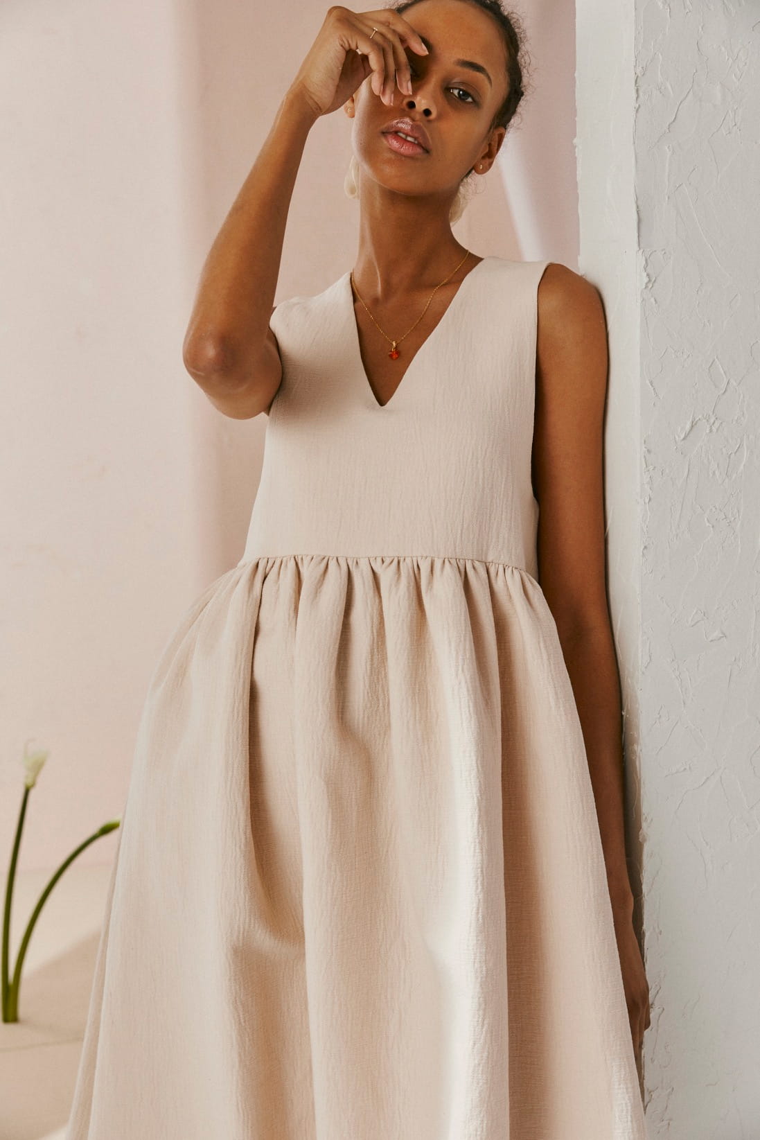 Wearing Noémiah's Marguerite Ring, a person in a sleeveless beige dress leans against a white textured wall. Their hand near their face draws attention to the daisy-inspired accessory. A delicate necklace complements the look, and the minimal interior setting further highlights the handmade jewelry with a single flower in the background.