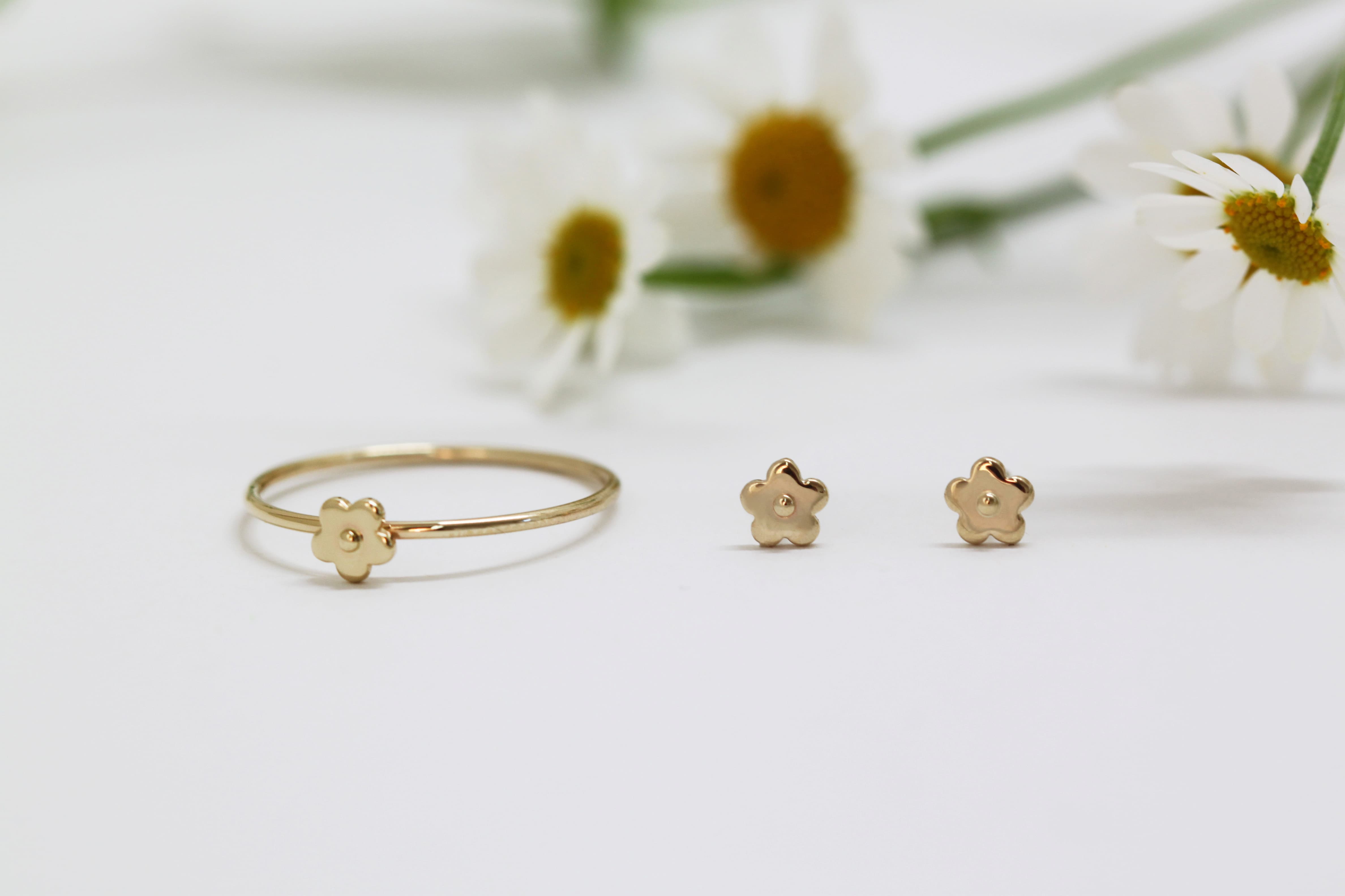 A delicate Marguerite Ring by Noémiah, crafted from solid gold, is accompanied by matching flower-shaped stud earrings on a white surface. In the background, blurred daisies enhance the handmade jewelry's simple elegance, adding a touch of charm to the setting.