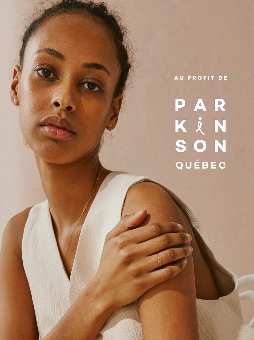 A woman in a white sleeveless top looks at the camera, gently touching her shoulder adorned with a Marguerite Ring by Noémiah. Text beside her reads Au profit de Parkinson Québec, indicating a charitable cause. The background is a soft beige color.