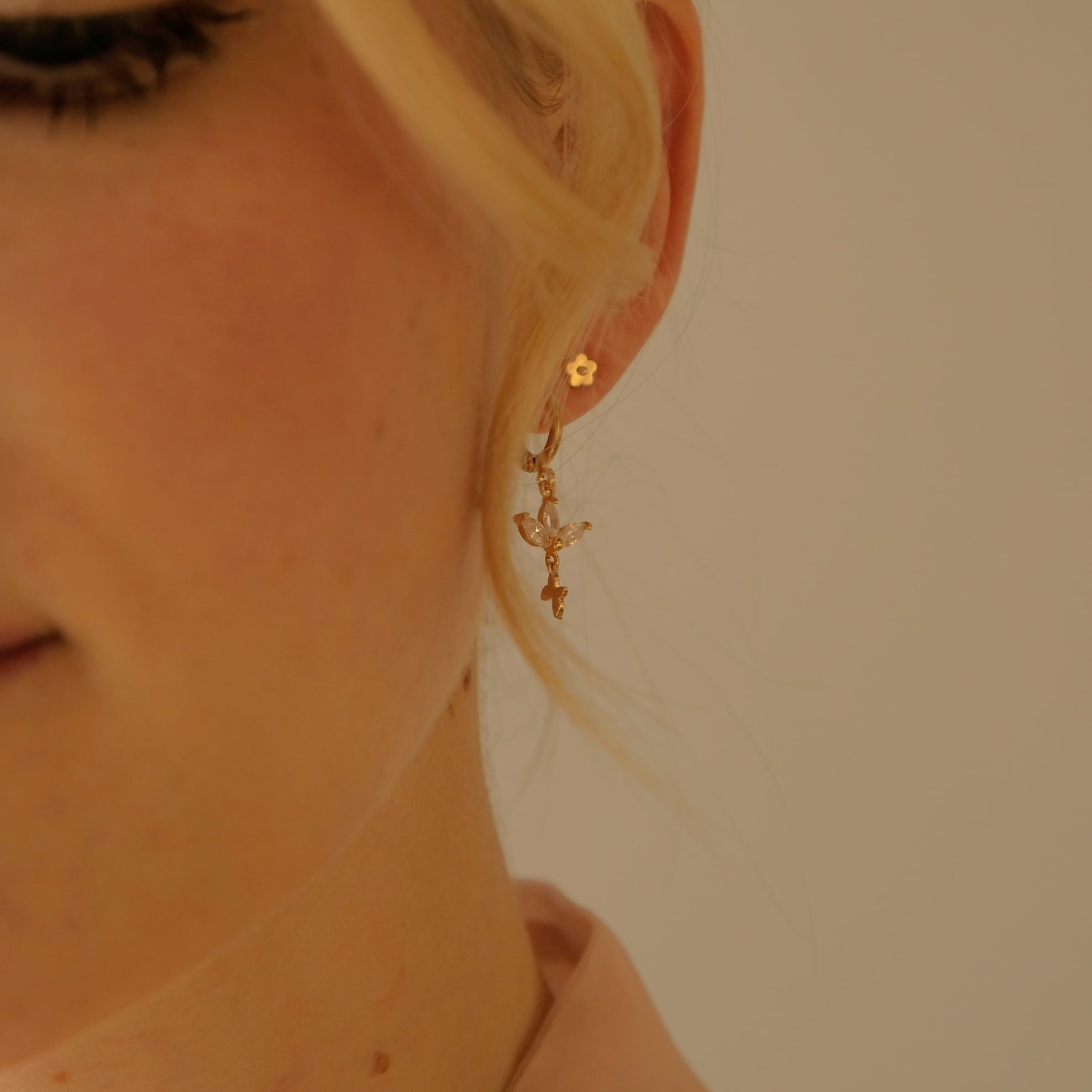 The scene captures a close-up of a person's ear decorated with the Marguerite Solid Gold Earrings by Noémiah. These handmade, 10-carat gold earrings boast an intricate floral design and a dangling cross. The individual's blonde hair is pulled back, highlighting the exquisite jewelry against a neutral background.