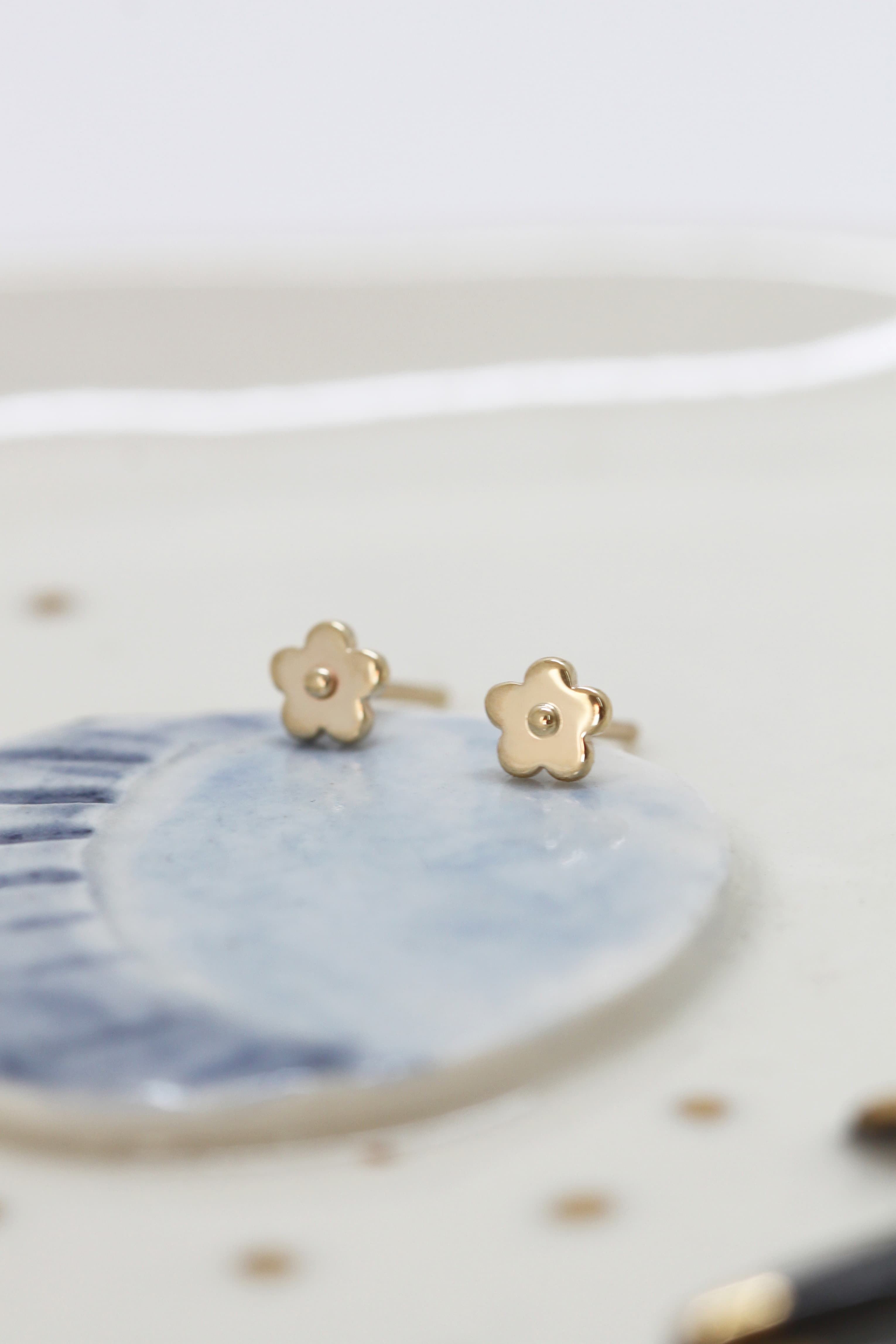 A pair of Marguerite Solid Gold Earrings by Noémiah, crafted from 10 carat gold, rests on a white and blue ceramic dish. The background is softly blurred, highlighting the delicate design of these handmade studs.