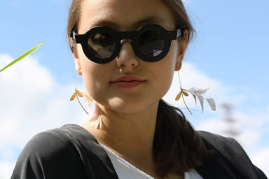 A woman wearing round black sunglasses and high-quality Noémiah Marie Earrings stands against a blue sky with white clouds. She has long hair and is dressed in a white shirt and dark outerwear, her earrings delicately crafted with gold-filled hooks.