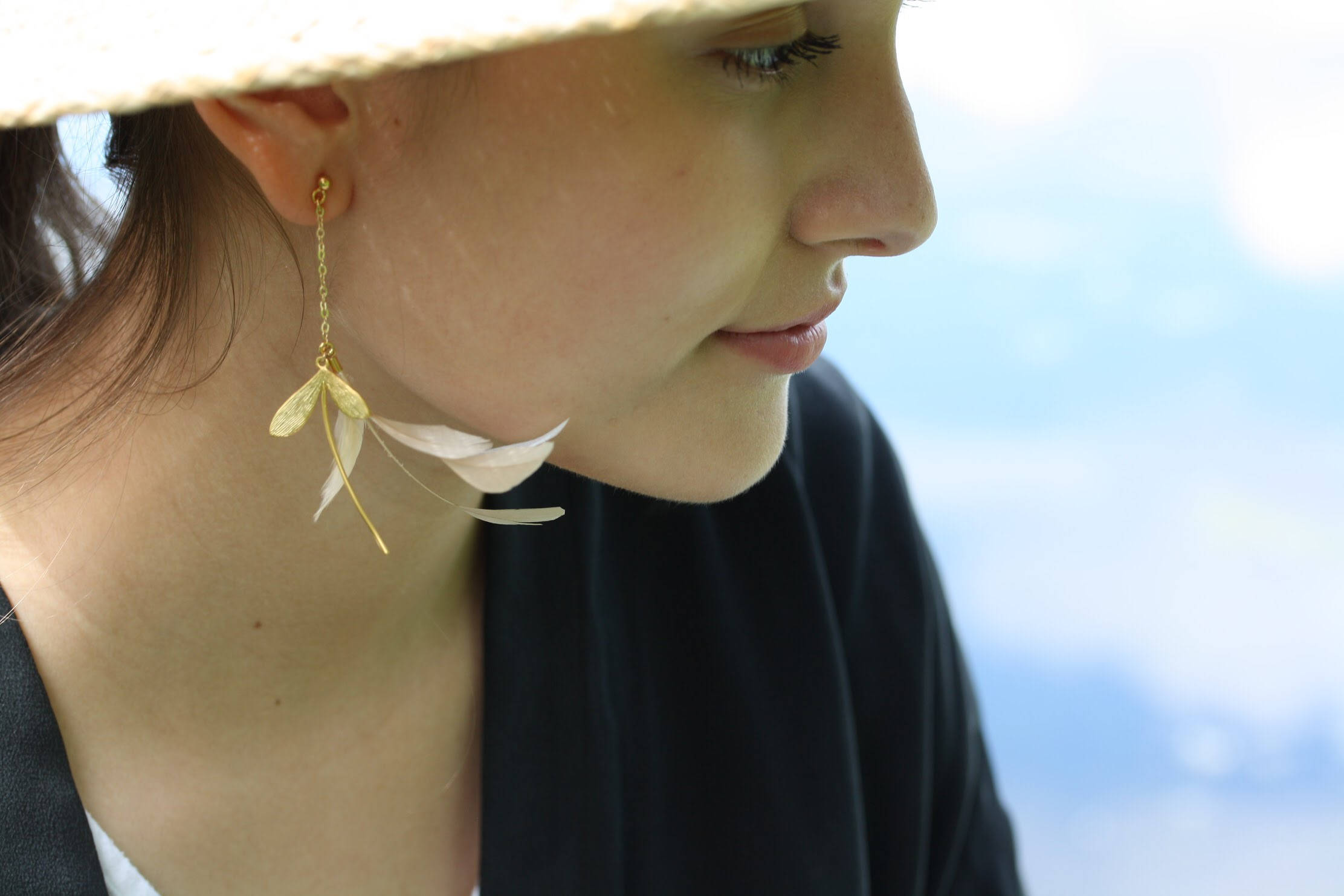 A woman in a straw hat is shown in profile, her serene expression complemented by the Marie Earrings from Noémiah, featuring lightweight gold-filled hooks and high-quality feathers. The softly blurred background suggests an outdoor setting.